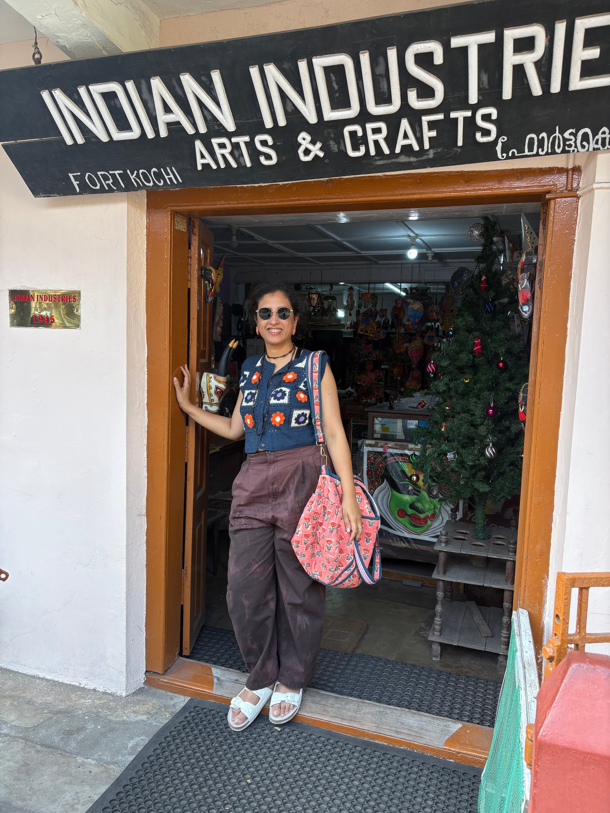 Woman standing outside a store named 'Indian Industries Arts & Crafts' with a Christmas tree inside.